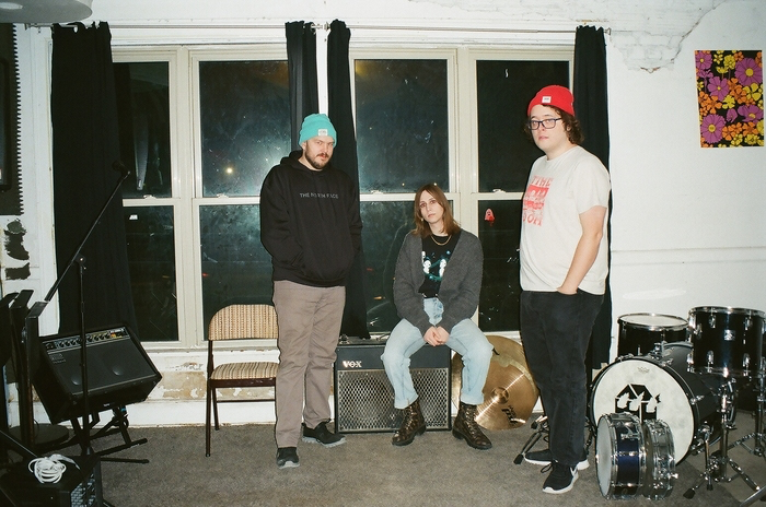 malamiko standing in their practice space, two members standing, one sitting on an amp. a broken down drumkit and amp are seen in the background